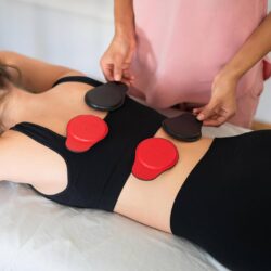 A patient is lying on their stomach on a white treatment table a patient lay down placing or adjusting several red and black magnetic pads on the patient's upper and lower back as part of a magnet therapy session