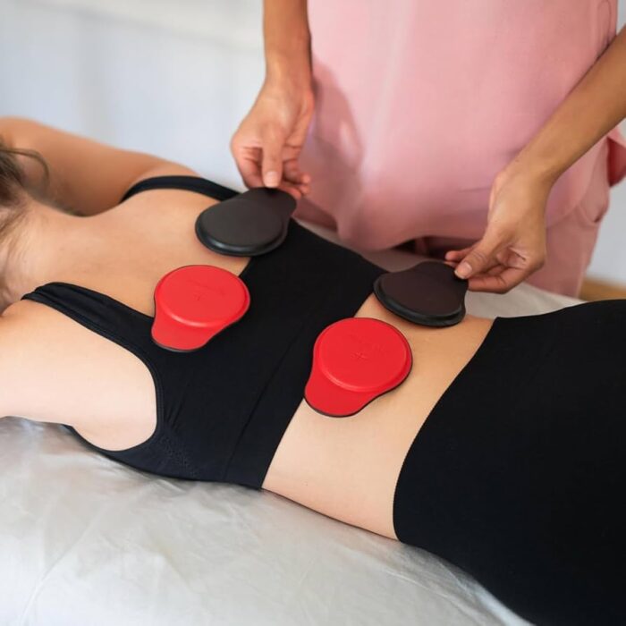 A patient is lying on their stomach on a white treatment table a patient lay down placing or adjusting several red and black magnetic pads on the patient's upper and lower back as part of a magnet therapy session